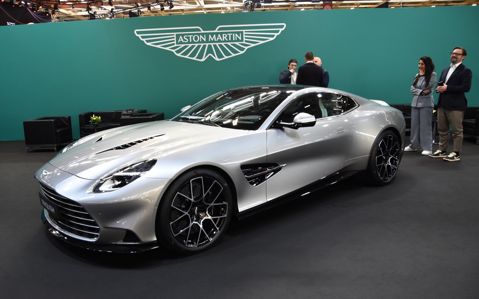 Silver Aston Martin Vanquish coupe on an indoor Aston Martin show stand, front three-quarter view, with the winged Aston Martin logo on the wall behind